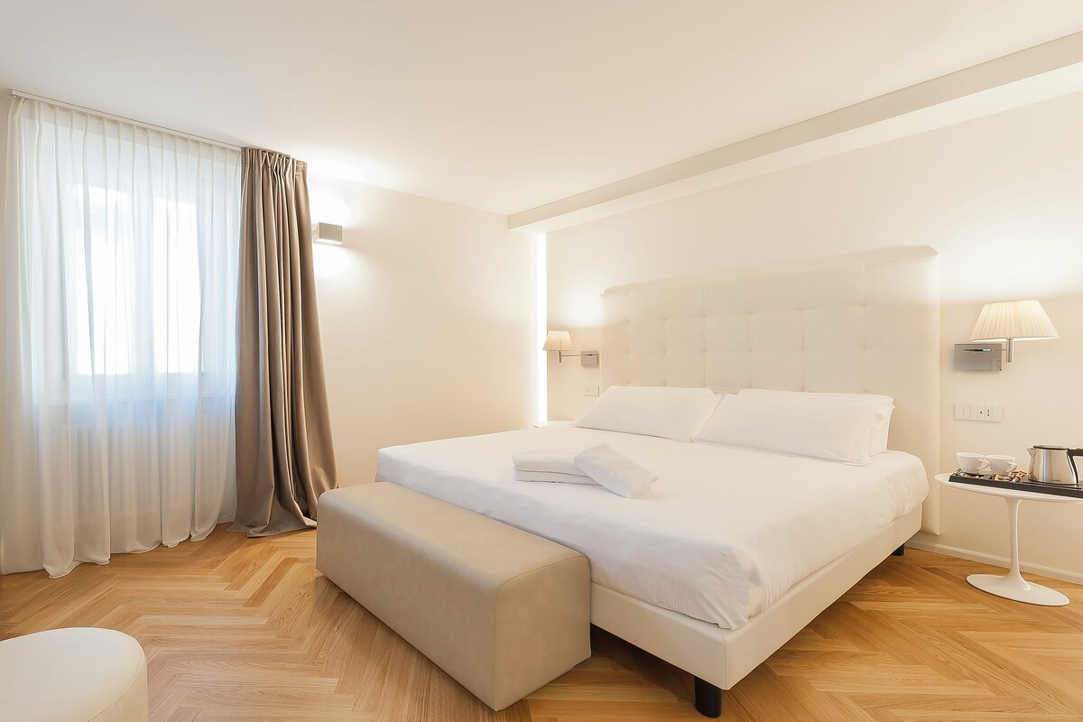 A hotel room with a large bed, a bedside table with a lamp, and a small table with cups and a kettle.