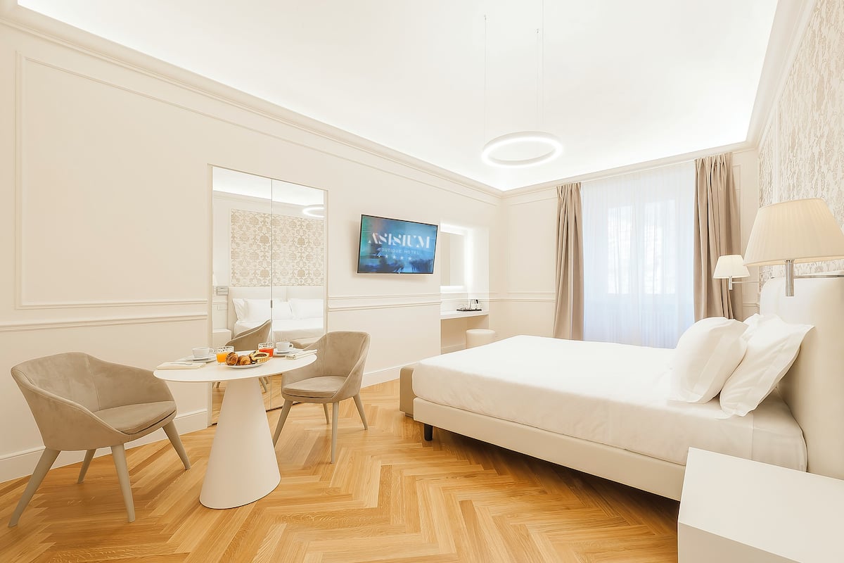 Ein Hotelzimmer mit einem Bett, zwei Sesseln, einem kleinen Tisch mit einem Getränk, einem Fernseher und einem Fenster mit Vorhängen.