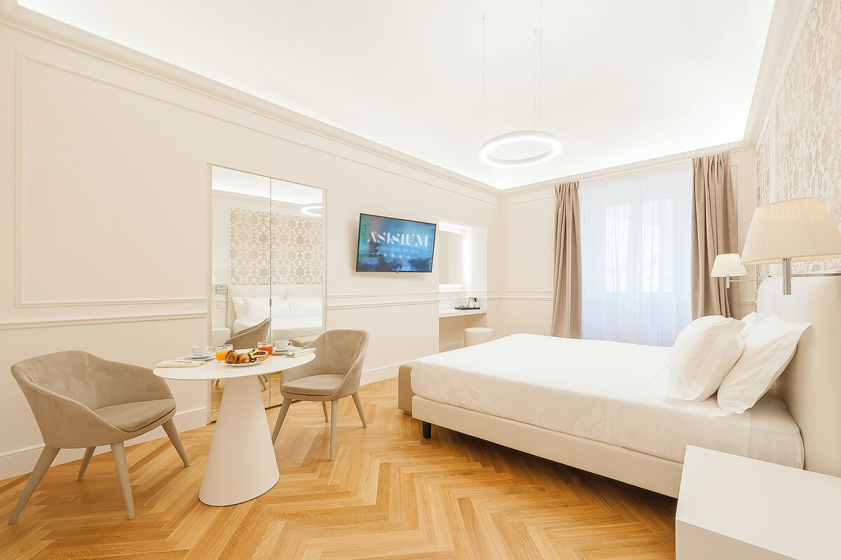 A hotel room with a bed, two chairs, a small table with a drink, a TV, and a window with curtains.