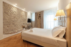 A modern hotel room with a stone accent wall, a flat-screen TV, a bed with white linens, and a large window with sheer curtains. - Asisium Boutique Hotel (Assisi)