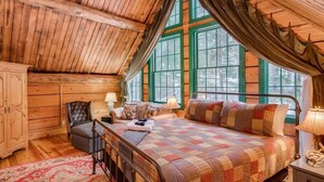 Une chambre confortable avec un plafond en bois, une grande fenêtre avec des volets verts, un lit recouvert d’une courtepointe, une chaise et une table de chevet avec une lampe.