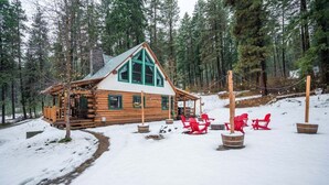 Une cabane en rondins dotée d’un porche couvert et de chaises Adirondack rouges, le tout situé dans un paysage forestier enneigé.