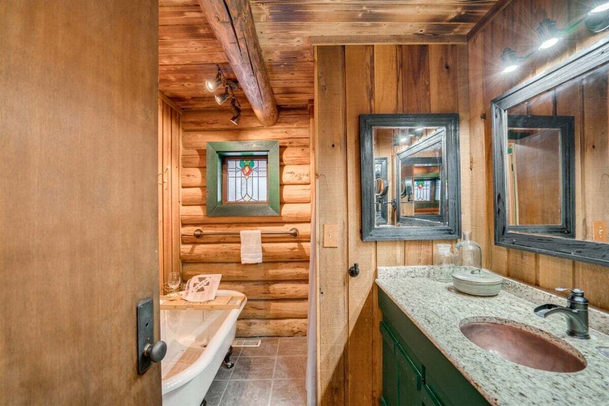 A wooden cabin-style bathroom with a wooden door, a sink with a granite countertop, a mirror, and a window with a stained glass design.