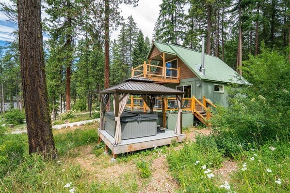 Una cabaña con terraza, jacuzzi y zona de estar exterior cubierta, rodeada de árboles.