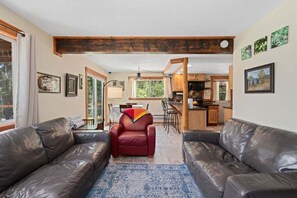 Una sala de estar con sofás de cuero, un sillón rojo, un tapete azul estampado, vigas de madera y una cocina al fondo.