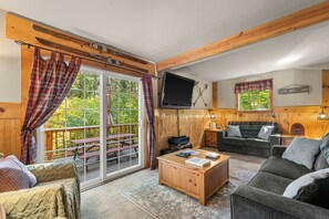 Een blokhutinterieur met een houten vloer, een bank, een salontafel, een televisie en een raam met gordijnen.
