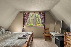 Een slaapkamer met een schuine plafond, een bed met een geborduurd sprei, een houten stoel, een bureau met een spiegel en een raam met ruitgordijnen.