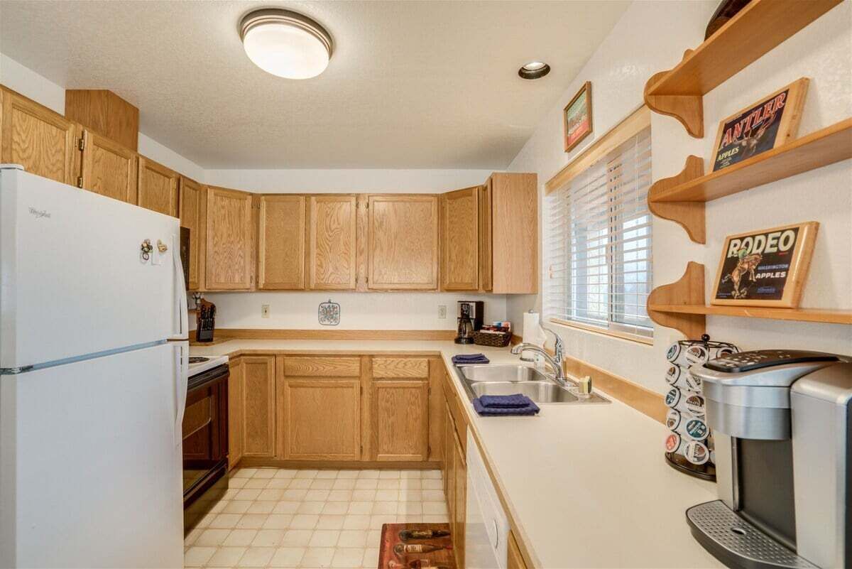 A kitchen with wooden cabinets, a refrigerator, a coffee maker, and a window with blinds.