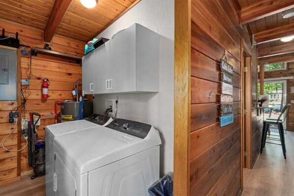 A laundry area with a washing machine, a dryer, and a fire extinguisher. There are wooden walls and a sign with room names. - Sunrise Lodge - Fetching Forest Cabin in the Sun! (Leavenworth)