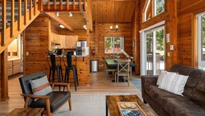 A wooden cabin interior with a dining area, kitchen, and a staircase leading to an upper level. - Sunrise Lodge - Fetching Forest Cabin in the Sun! (Leavenworth)
