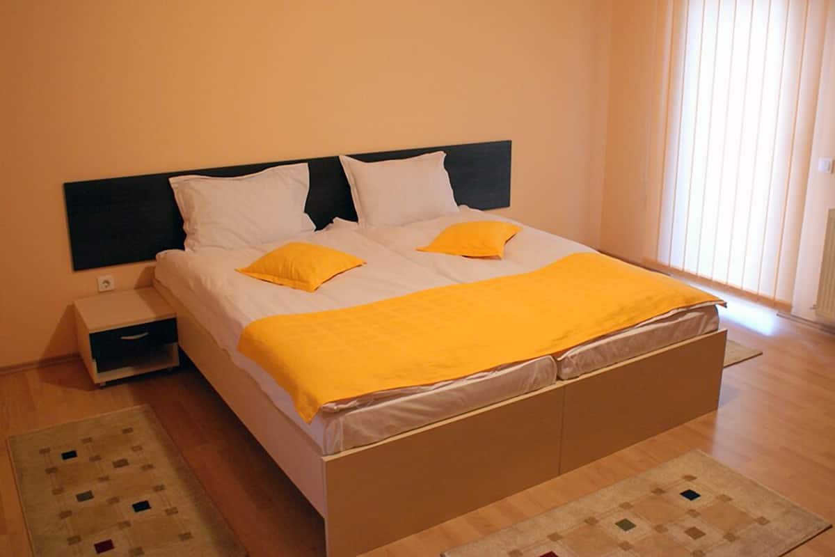 Uma cama de solteiro com cabeceira preta, roupa de cama branca e duas almofadas amarelas. Há uma mesa de cabeceira com gaveta, uma janela com persianas e um tapete estampado no chão.