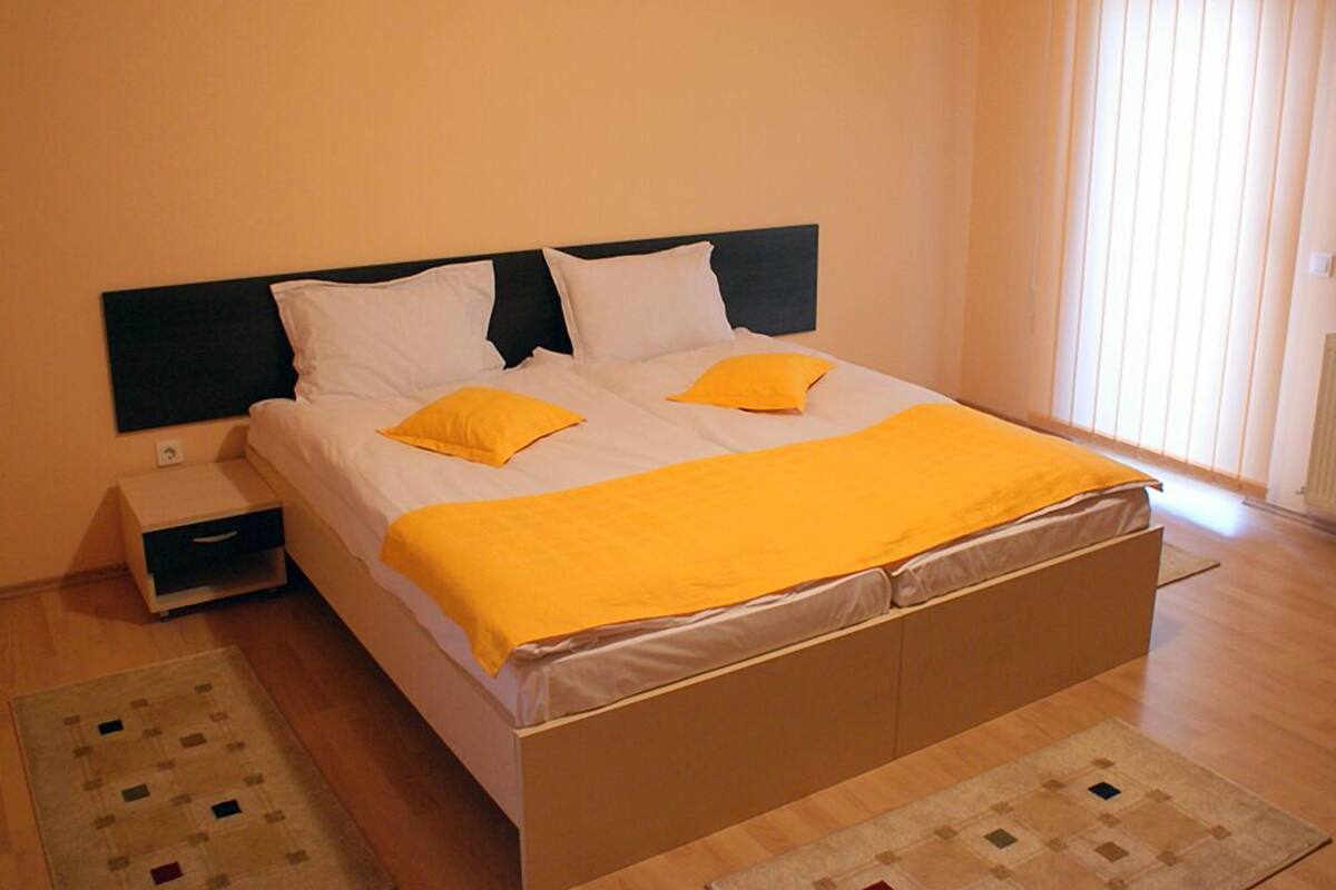 A single bed with a black headboard, white bedding, and two yellow pillows. There is a nightstand with a drawer, a window with blinds, and a patterned rug on the floor.