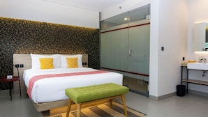 A modern hotel room with a large bed, a green bench, a glass-enclosed wardrobe, and a bathroom with a sink and mirror. - Qubika Boutique Hotel (Gading Serpong)