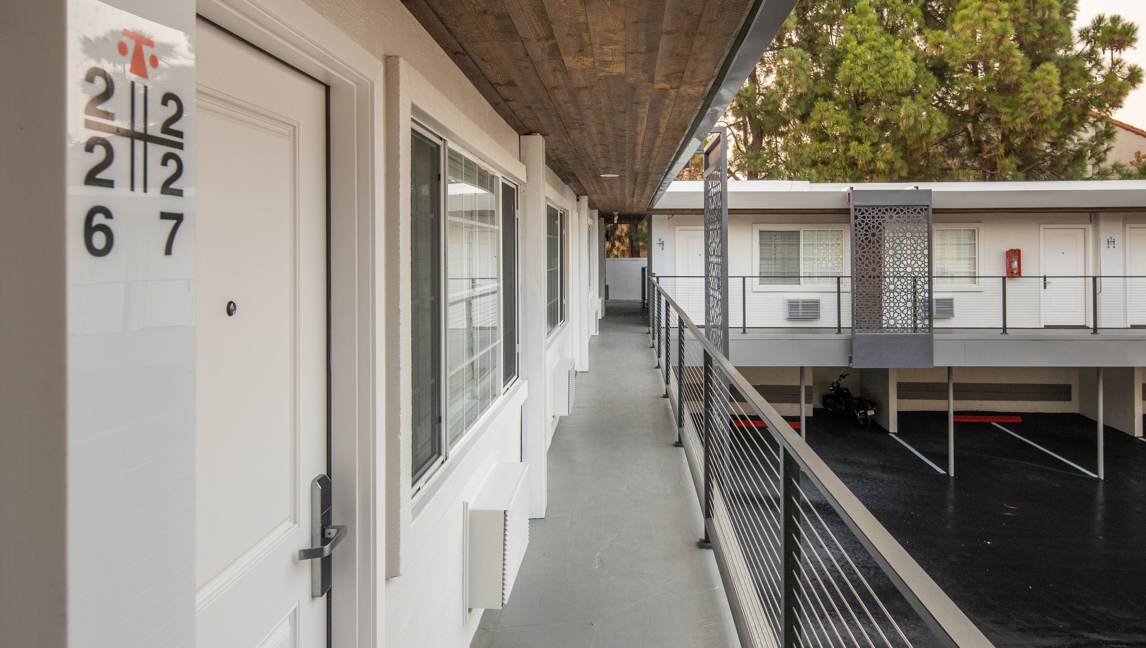 A corridor with numbered doors, a covered walkway, and a parking area with a motorcycle.