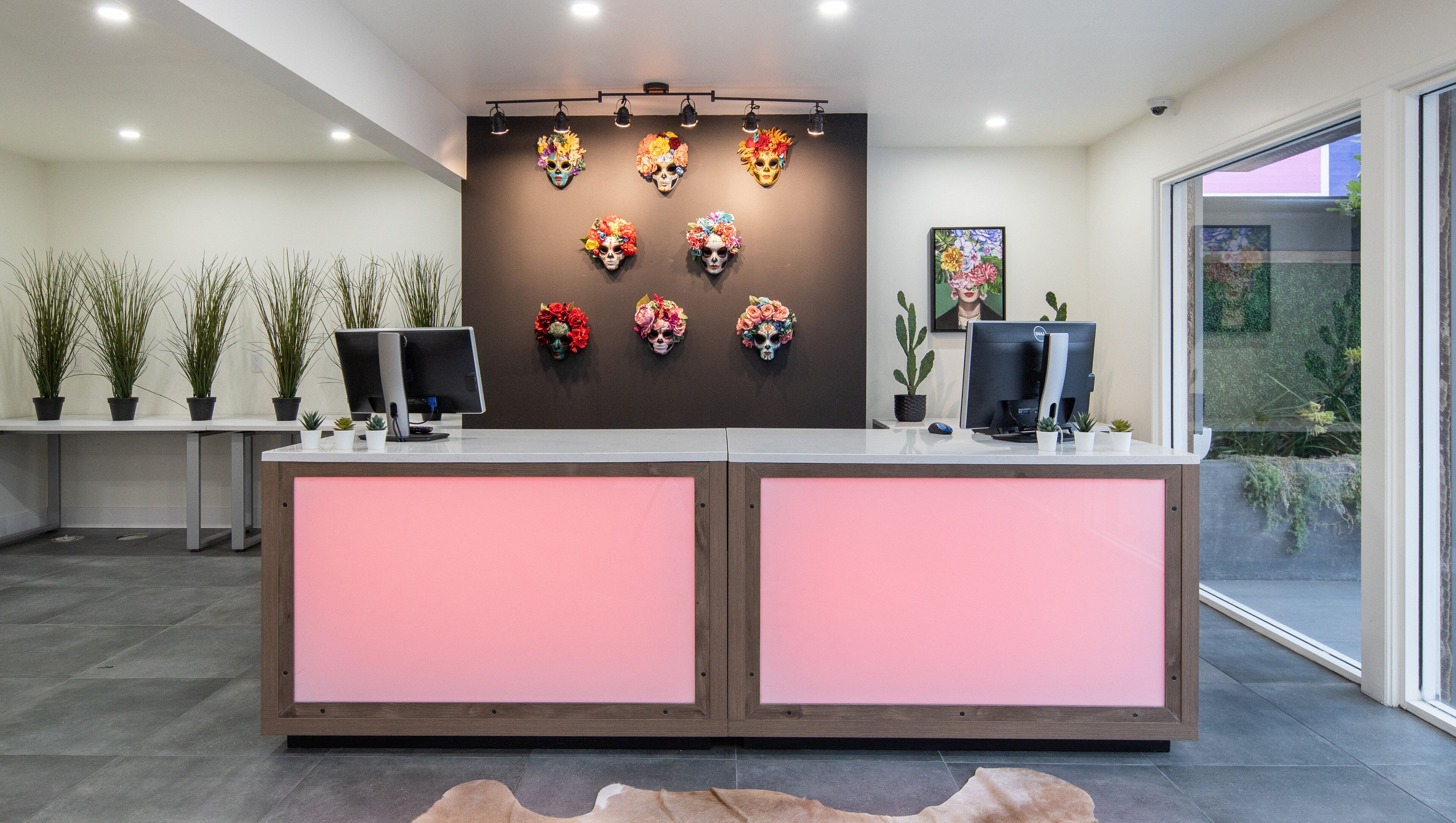 A reception desk with two computer monitors, a wall-mounted light fixture, and a row of potted plants.