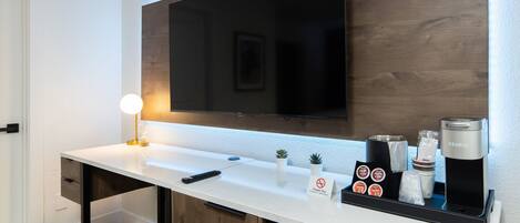 A modern hotel bathroom with a white countertop, a mounted television, a Keurig coffee machine, and a sign prohibiting smoking.