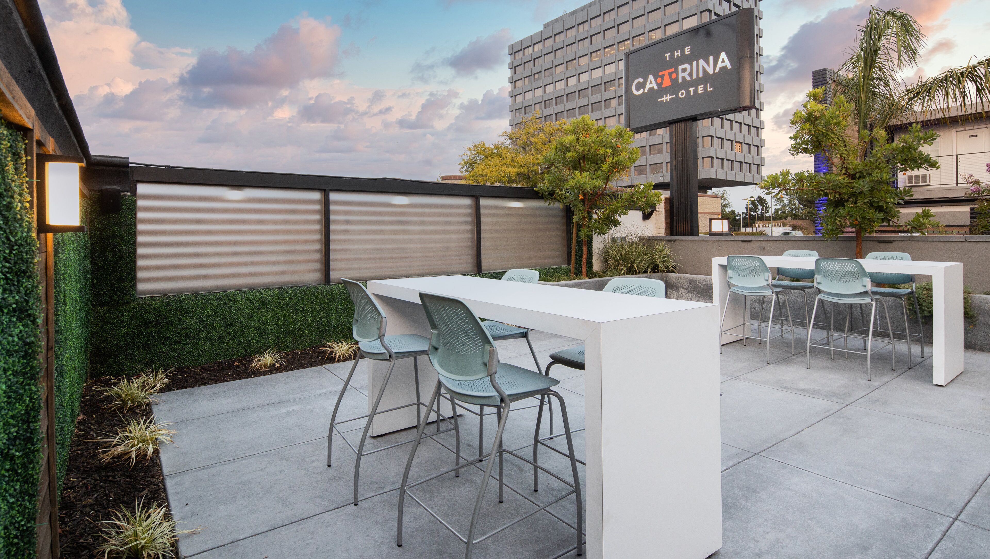 An outdoor patio area with a white table and chairs, a sign for 'The Carrina Hotel', and a modern building in the background.