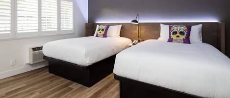 A hotel room with two beds, a bedside table with a lamp, and a window with blinds.