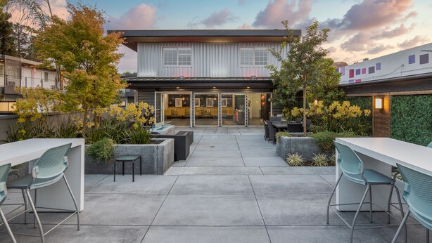 An outdoor patio area with a modern building in the background, featuring a glass door entrance, outdoor seating, and a variety of plants. - The Catrina Hotel (San Mateo)
