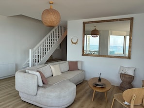Sala de estar con sofá blanco, mesa redonda de madera, silla de mimbre, una escalera y un espejo grande que refleja la vista de un balcón.