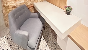 Une pièce moderne avec un mur en pierre, un fauteuil à revêtement gris, un bureau blanc et une petite plante en pot.
