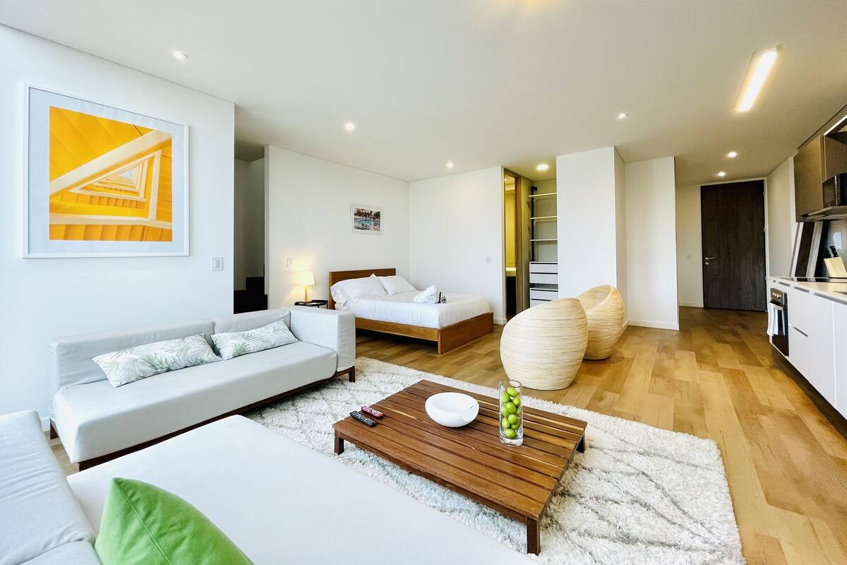 A modern living room with a sofa, a coffee table, and a bed. There is a staircase leading to a loft area.