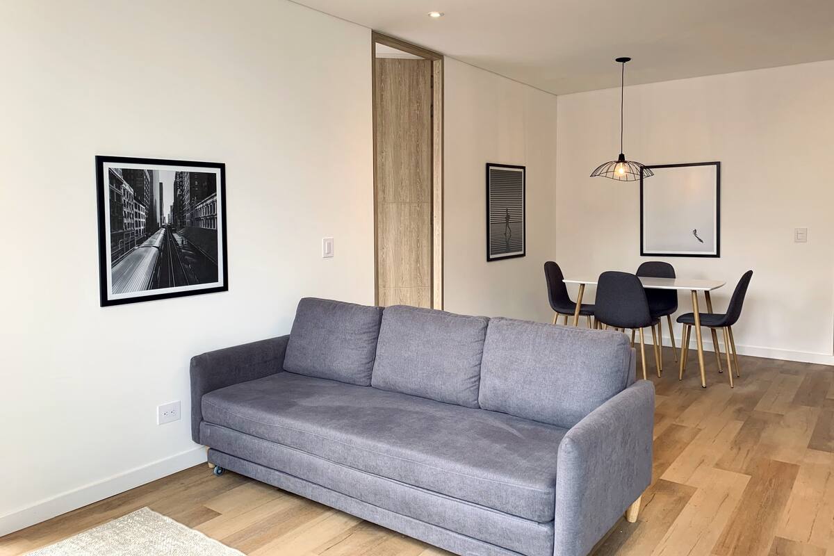 A modern living room with a grey sofa, a dining area with a white table and black chairs, and a wooden floor.