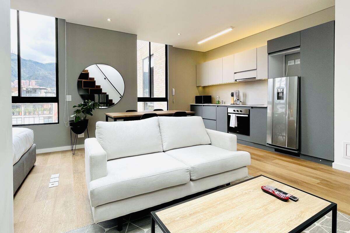 A modern apartment with a living area featuring a sofa, a coffee table, and a dining table. The kitchen includes a refrigerator, stove, and cabinets.