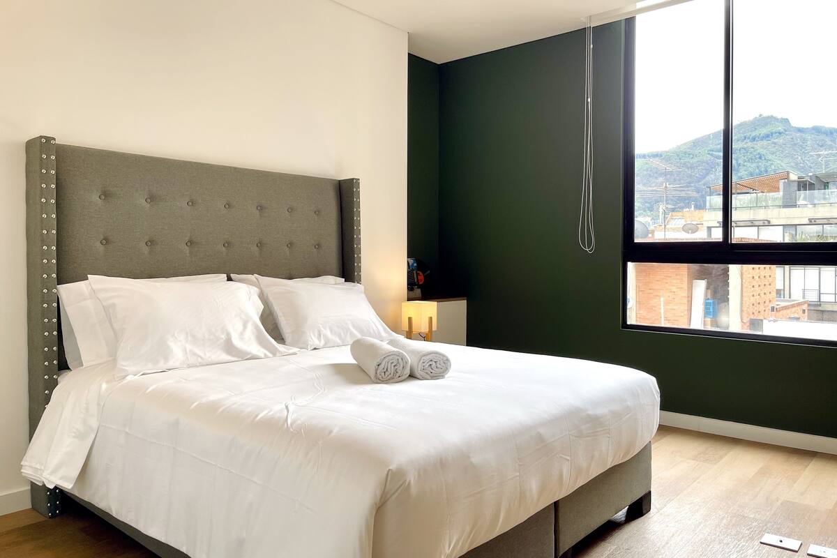 A neatly made bed with white linens and a grey headboard, a bedside lamp, and a view of a cityscape through a large window.
