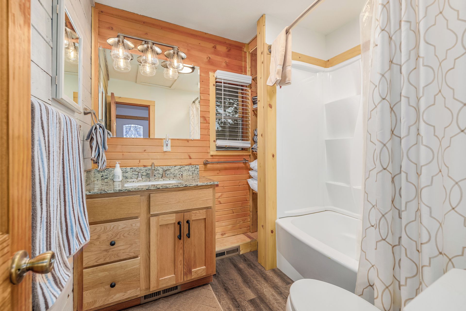 A bathroom with wooden paneling, a sink with a mirror, a shower with a patterned curtain, and a towel hanging on the door.