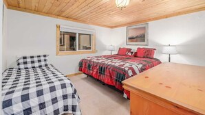 A bedroom with a wooden ceiling, a bed with a red and black checkered bedspread, a smaller bed with a black and white checkered bedspread, a window, and a painting on the wall. - Cabin 1, Aspen Leaf Lodges, Near Grand Lake (Grand Lake)