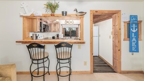 A compact kitchen with wooden cabinets, a built-in microwave, and a window above the sink. There are two metal chairs at a small table. A wooden door leads to another room, and a sign on the wall reads "LIFE IS BETTER AT THE LAKE". - Cabin 1, Aspen Leaf Lodges, Near Grand Lake (Grand Lake)
