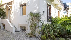 Un edificio blanco con una escalera que lleva a una puerta, un pequeño jardín con plantas y un patio empedrado.