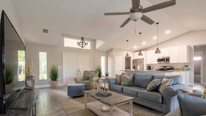 A spacious living room with a large grey sofa, a coffee table, and a TV. There is a ceiling fan, pendant lights, and a kitchen in the background. - Luxury Cape Coral Retreat Pool, Hot Tub, Beautiful Outdoor Oasis & Pet Friendly (Cape Coral)