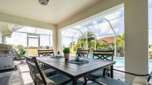 Un patio techado al aire libre con una mesa y sillas, una parrilla y vistas a una piscina y vegetación.