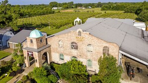 Ein aus Ziegeln errichtetes Gebäude mit einer Kuppel und einem Bogenportal, das „Haak Winery“ genannt wird, umgeben von einem gepflegten Garten und einem Friedhof im Hintergrund.