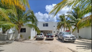 A parking area with several cars, palm trees, and a building with a flat roof and windows. - Casa Sara  -  Yucatan Home Rentals (Chicxulub Puerto)