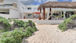 A beachside resort with a thatched-roof structure, outdoor seating, and a sandy pathway leading to a building with large windows. - Casa Sara  -  Yucatan Home Rentals (Chicxulub Puerto)