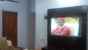 A room with a wooden door, a striped sofa, and a television mounted on the wall displaying a person. - JiL Service Flat (Ibadan)