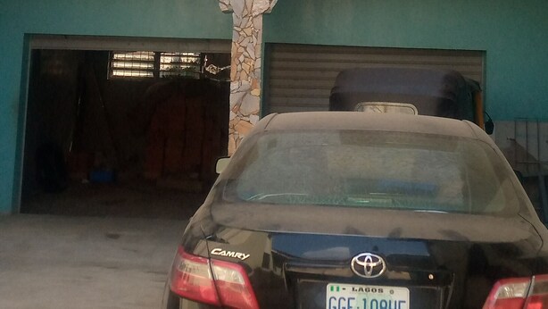 A black Toyota Camry parked in front of a building with a stone facade and two garage doors. - JiL Service Flat (Ibadan)