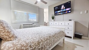 Une chambre à coucher moderne avec un lit, un téléviseur fixé au mur, une commode et une fenêtre.
