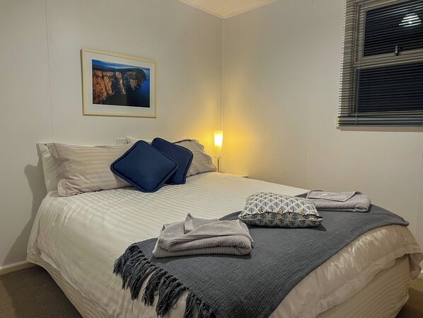 Una cama bien hecha con una colcha estampada, almohadas y una toalla doblada. Un cuadro enmarcado cuelga de la pared sobre la cama.