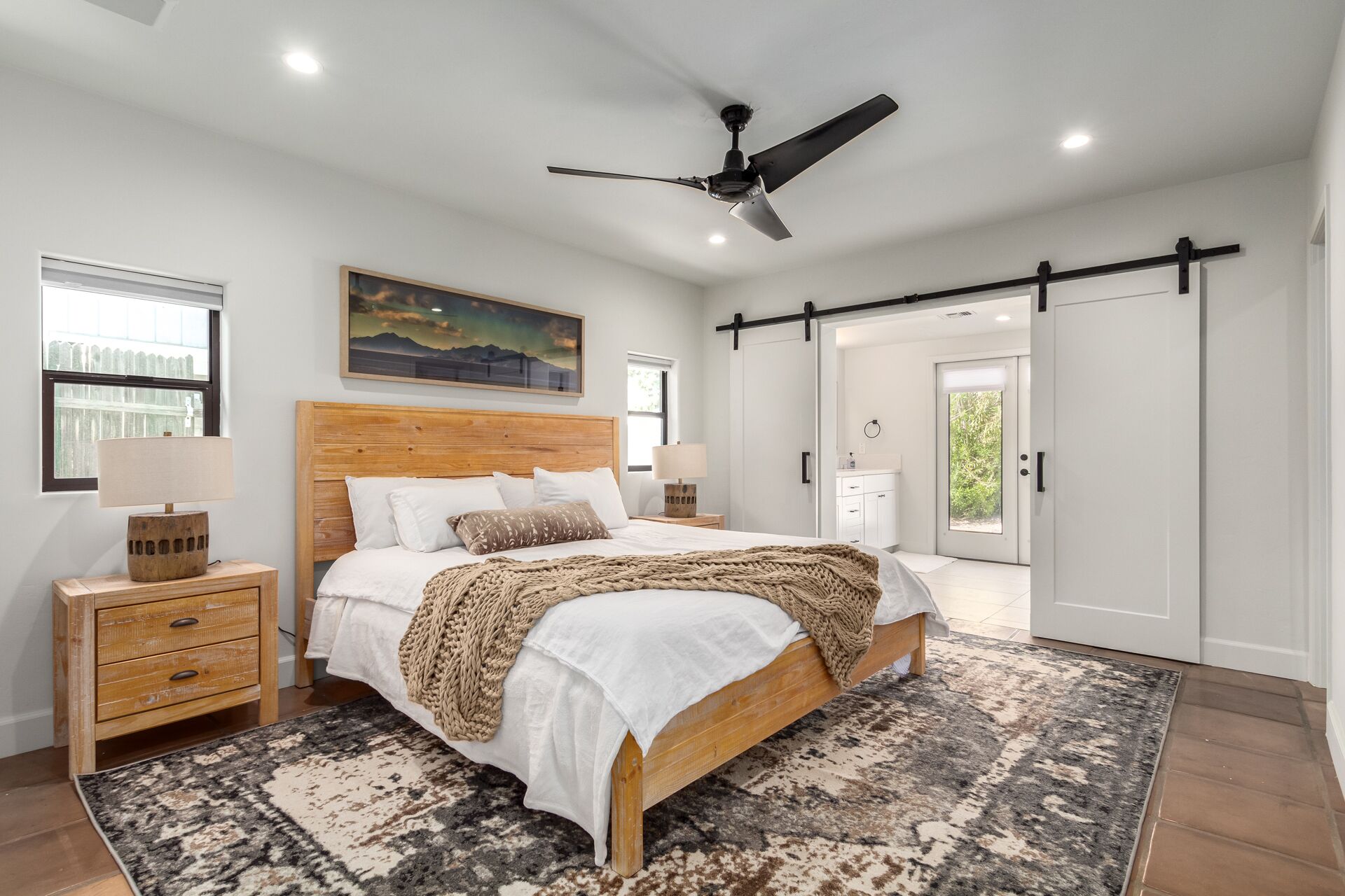 Un dormitorio con una cama de madera, un ventilador de techo, un ventanal grande y una puerta corredera que da a un baño.