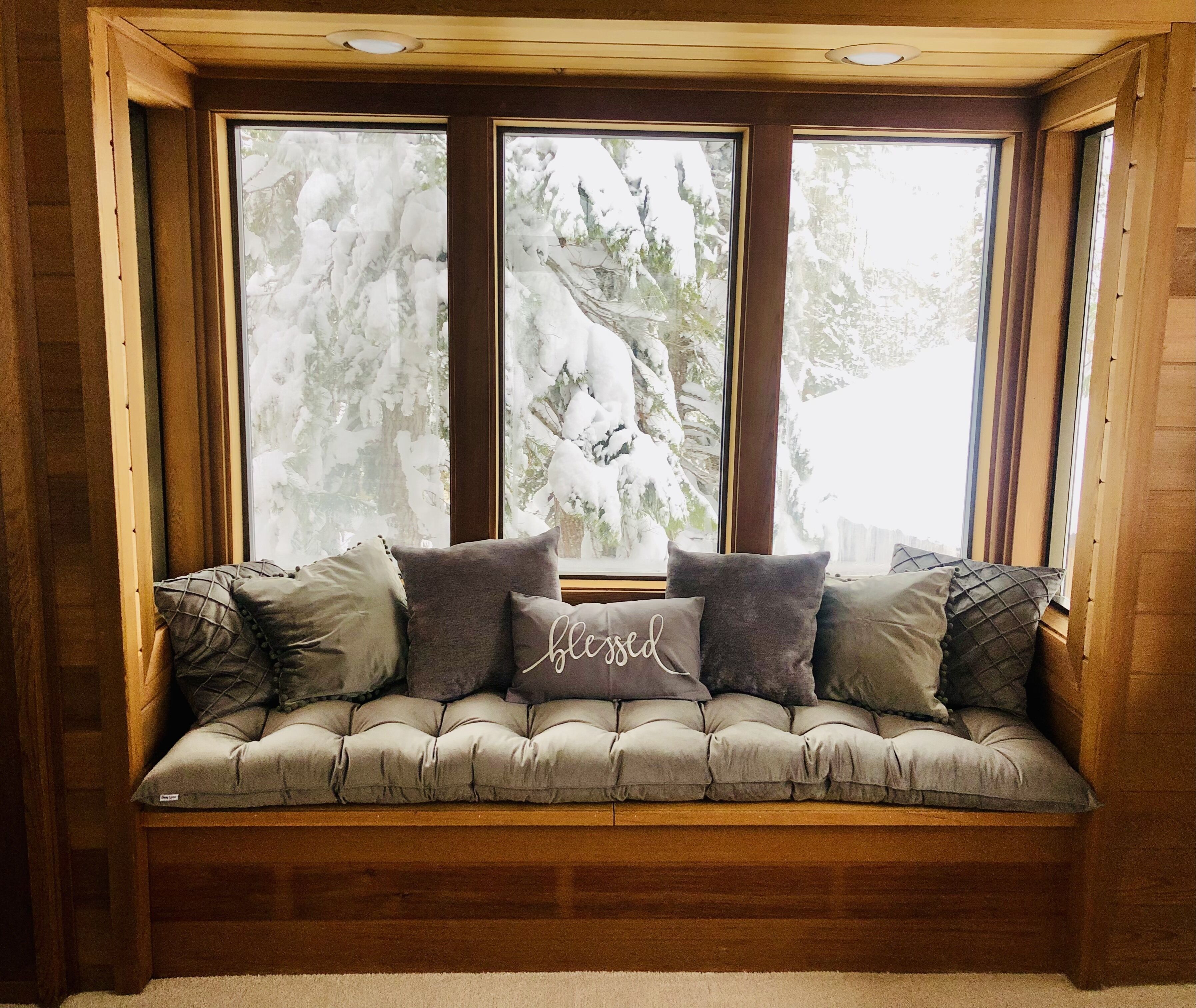 Un banc en bois confortable, doté d’un siège rembourré et de coussins, placé sous une grande fenêtre offrant une vue sur des arbres couverts de neige.