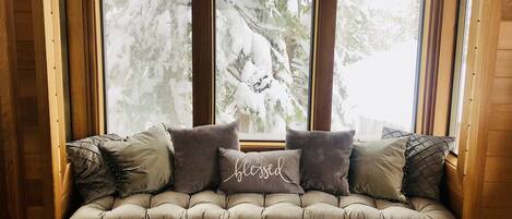Un banc en bois confortable, doté d’un siège rembourré et de coussins, placé sous une grande fenêtre offrant une vue sur des arbres couverts de neige.