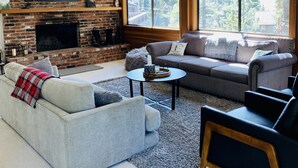 A cozy living room with a fireplace, a large sofa, a TV mounted on a brick wall, and large windows offering a view of trees. - Our Lake Tahoe H💙ME<br> (Tahoe City)