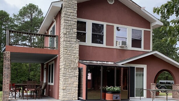 A two-story house with a stone chimney, a covered patio with outdoor furniture, and a garage with a vehicle. - Beautiful home and/or garage apartment, minutes from Oaklawn Park. (Hot Springs)