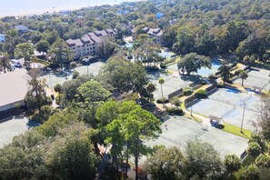 An aerial view of a residential area with multiple tennis courts, a swimming pool, and surrounding trees.