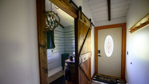 Un pasillo interior de madera con alfombra a cuadros, un barril y una puerta con un ventanal ovalado.