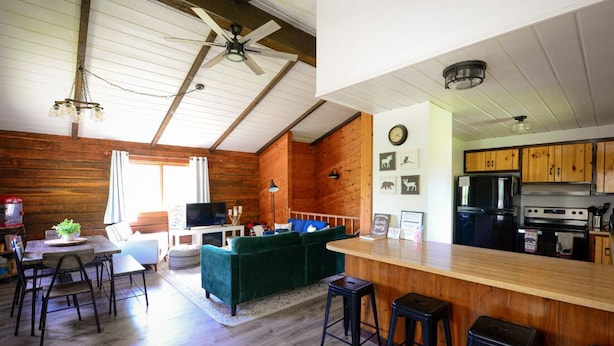 Un acogedor interior de cabaña con comedor de madera, un sofá verde, una barra con taburetes y una cocina con armarios y electrodomésticos de madera.